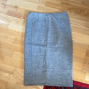 Grå kjol i syntetmaterial - Snygg grå kjol i syntetmaterial med en tight passform. Kjolen har en klassisk design och når till knäna, vilket ger en stilren look. Perfekt för både vardag och mer uppklädda tillfällen.