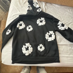 Svart hoodie med vita blommor från Denim Tears - Säljer en svart hoodie från Denim Tears med vita blommönster. Den har en stor ficka framtill och en bekväm huva. Perfekt för en avslappnad stil.