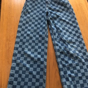 Blå rutiga jeans med vida ben - Snygga blå jeans med ett rutigt mönster och vida ben. De är högmidjade och perfekta för en trendig look. Tillverkade i Bangladesh, dessa jeans är både stiliga och bekväma.