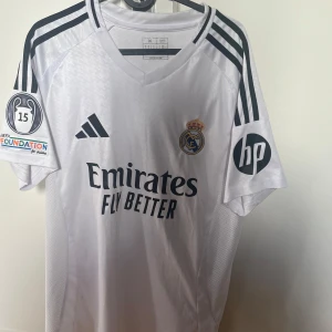 Real Madrid fotbollströja - Säljer en vit Real Madrid fotbollströja från Adidas med korta ärmar. Tröjan har svarta detaljer och sponsorloggor på ärmarna. Namnet 'Mbappé' och nummer 9 är tryckt på baksidan. Perfekt för fotbollsfans! Köpt på Bernabeu i sportshoppen. Player edition 