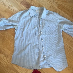 Blå- och vitrandig skjorta - Säljer en stilren blå- och vitrandig skjorta med lång ärm och klassisk krage. Skjortan har knappar framtill och en bröstficka. Perfekt för en avslappnad men ändå uppklädd look.