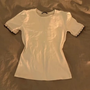 Vit kortärmad topp med broderade detaljer - Säljer en stilren vit topp med korta ärmar och unika broderade detaljer vid ärmsluten. Perfekt för en enkel men elegant look. Passar bra till både jeans och kjol.