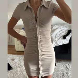 Beige kortklänning med knappar - Säljer en stilren beige kortklänning med knappar hela vägen ner. Klänningen har korta ärmar och en klassisk krage. Stretchig!