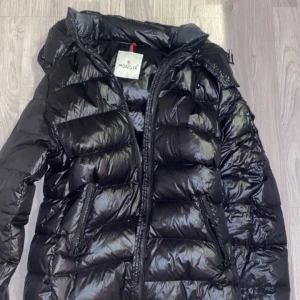 Svart dunjacka från Moncler - Säljer en snygg svart dunjacka från Moncler. Jackan har en glansig finish och är utrustad med dragkedja och praktiska fickor. Perfekt för kyliga dagar när du vill hålla stilen.  Säljer jackan för jag trodde det var för herrar. Mötts upp helst.