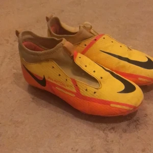 Gula och orange fotbollsskor från Nike - Säljer ett par Nike Phantom fotbollsskor med svarta och oranga detaljer. Skorna har en unik och modern design med snöring och är perfekta för fotbollsplanen och för snabba rörelser. Skorna har en svart Nike-logga och är designade för optimal prestanda på planen. De har en slitstark sula med dobbar för bra grepp. Fotbollsskorna är storlek 36 men rekommenderar att inte köpa om man har exakt 36 så skorna är mindre i passform. Perfekt för dig som vill sticka ut!