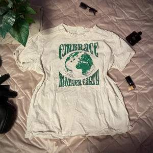 Vit t-shirt med tryck från Montalami - Säljer en vit t-shirt från Montalami med ett grönt tryck som säger 'Embrace Mother Earth'. T-shirten har korta ärmar och är perfekt för en avslappnad stil. Materialet är mjukt och bekvämt. Har två små nästan osynliga fläckar på baksidan men syns knappt. 