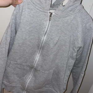 Grå hoodie med dragkedja från 157 - Säljer en grå hoodie från 157 med dragkedja och huva. Tröjan har långa ärmar och praktiska fickor framtill. Perfekt för en avslappnad stil. Hör av dig vid intresse! Frakt tillkommer.