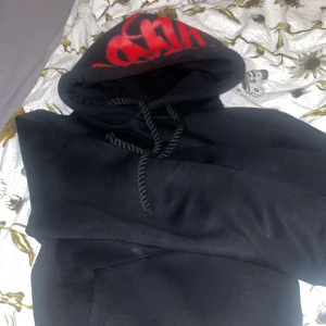 Svart hoodie med röd text - Säljer en svart hoodie med en stor röd text på huvan. Den har en klassisk design med dragsko i huvan och en stor ficka framtill. Perfekt för en avslappnad stil.