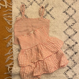 Rosa rutig topp och kjol - Söt rosa rutig set med en smockad topp och matchande kjol. Toppen har smala axelband och kjolen har volanger för en lekfull look. Perfekt för varma sommardagar!