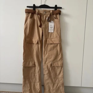 Beige cargobyxor från Bershka - Säljer ett par beige cargobyxor från Bershka med raka ben och flera praktiska fickor. Byxorna har en medföljande brunt bälte och är perfekta för en avslappnad stil. De är tillverkade i ett slitstarkt material och har en klassisk cargodesign.
