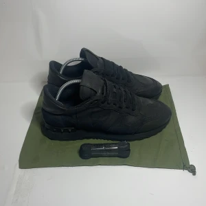 Valentino rockrunner - Snygga svarta sneakers från Valentino med ikoniska nitar på hälen. Storlek 39.5 men passar 40 Skorna har en stilren design med snörning och är tillverkade i mocka. Perfekta för en trendig look.