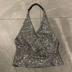 Glittrig silver topp från Zara - Säljer en glittrig silver topp från Zara med paljetter. Toppen har en omlott-design och halterneck. Perfekt till fest! Strl L men passar även en M