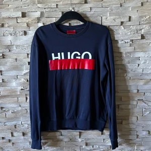 Mörkblå sweatshirt från Hugo - Snygg mörkblå sweatshirt från Hugo med vit logotyp och röd blank detalj på bröstet. Tröjan har rund hals och långa ärmar, perfekt för dig som gillar stilrena och trendiga plagg. Passar dig som vill ha en enkel men cool look.