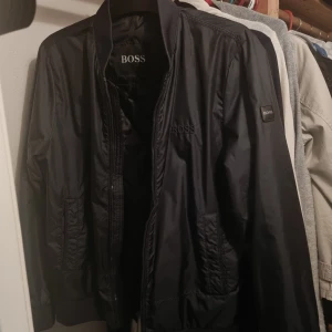 Svart bomberjacka från BOSS - Snygg svart bomberjacka från BOSS med quiltat foder och ribbade muddar. Jackan har dragkedja framtill, två sidofickor och BOSS-logga på bröstet och ärmen. Perfekt för dig som gillar stilren design och vill ha en jacka som funkar till många outfits.