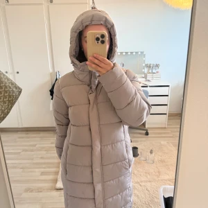 Jacka puffer - Säljer en beige lång pufferjacka med huva och dragkedja framtill. Jackan har ribbade muddar, fleecefodrade fickor och snygga detaljer med knappar och snörning vid huvan. Perfekt för kalla dagar när man vill vara både varm och stilren. Passar S-M