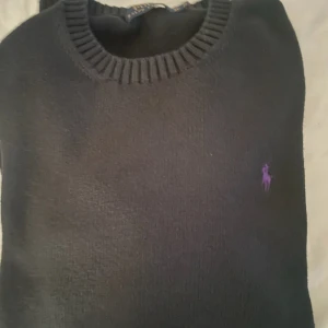 Svart stickad tröja från Polo Ralph Lauren - Klassisk svart stickad tröja från Polo Ralph Lauren i storlek XL. Tröjan har rund halsringning med bred ribbad kant och en lila broderad logga på bröstet. Perfekt för dig som gillar stilren och tidlös design.