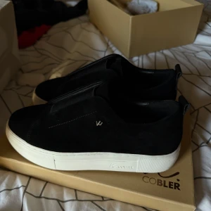 Svarta sneakers från K.Cobler i mocka - Stilrena svarta sneakers från K.Cobler med ovandel i mjuk mocka och bred resår över vristen. Skorna har vit platt sula och diskret logga på sidan. Perfekta för en clean och modern look. Helt nya och endast testade då de va för små