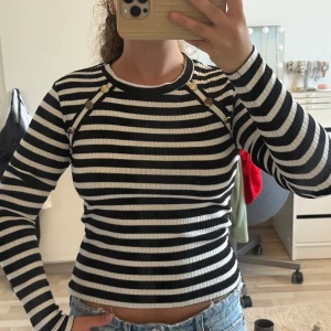 Långärmad tröja, ribbad - Säljer en svartvit randig ribbad tröja med lång ärm och snygga guldfärgade knappar vid axlarna. Helt oanvänd