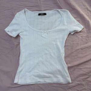 Supersöt t-shirt - Supersöt vit t-shirt med lite mönstrat tyg och en rosett uppe vid kragen! Den är endast andvänd 1-2 gånger och är ifrån bikbok!