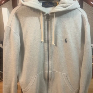 Polo Ralph Lauren Tröja Grå - Snygg grå hoodie från Polo Ralph Lauren med klassisk blå logga på bröstet. Tröjan har huva med snörning, dragkedja framtill och två fickor!Gamla modellen, säljs knappt längre och sällsynt. Passar bra till allt, tveka inte att höra av dig:)
