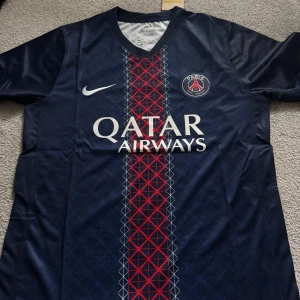 PSG Hemma ställ 25/26 - Paris Saint-Germain fotbollströja från Nike i marinblått med rött och vitt geometriskt mönster längs mitten. Tröjan har korta ärmar, V-ringad krage och PSG-logga på bröstet. Materialet är lätt och andas, Dri-FIT-teknologi. Qatar Airways tryck på framsidan.