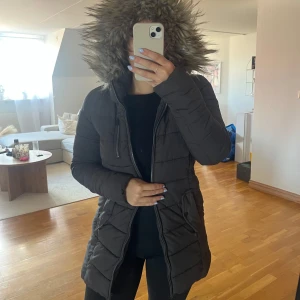 Svart pufferjacka med fuskpäls från ONLY - Säljer en svart pufferjacka från ONLY i storlek XS. Jackan har en stor huva med fuskpäls, dragkedja framtill och praktiska fickor. Perfekt för kalla dagar med sin långa modell och quiltade design. Snygg och bekväm att bära hela vintern.