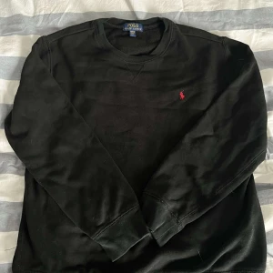 Svart sweatshirt från Polo Ralph Lauren - Svart sweatshirt från Polo Ralph Lauren med klassisk rund hals och röd broderad logga på bröstet. Tröjan har långa ärmar och är tillverkad i mjuk bomull, perfekt för en clean och stilren look. Pris kan diskuteras 