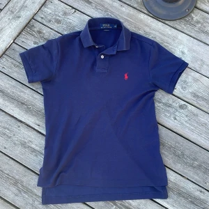 Marinblå pikétröja från Ralph Lauren - Snygg marinblå pikétröja från Polo Ralph Lauren i bomull. Klassisk krage, korta ärmar och två knappar framtill. Ikoniska röda polospelaren broderad på bröstet. Custom fit-modell som sitter riktigt bra och passar till allt. Priset ej hugget i sten! Hör av er vid funderingar!🙌