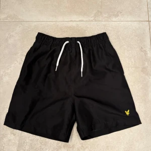 Svarta Lyle & Scott badshorts med snörning - Svarta badshorts från Lyle & Scott med innerbyxa i nät. Snörning i midjan och resår. Sidofickor och en bakficka med lock och kardborre. Passar från 10-11 år. (Mäter 38 cm i midjan vid utdragen resår, från kant till kant, en sida). 