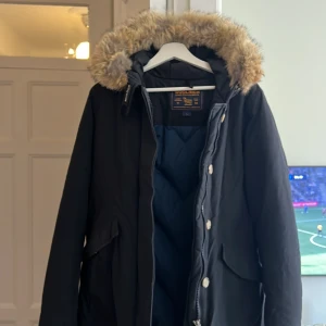 Svart parkas från Woolrich med pälskrage - Säljer en svart parkas från Woolrich med stor fluffig pälskrage i beige och grå toner. Jackan har dragkedja och knappar framtill, stora fickor och är fodrad för extra värme. Perfekt för kalla dagar och riktigt snygg streetstil.   Passar storlek 38. Avtag bar päls