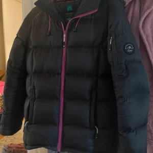 Svart dunjacka från Tuxer med rosa detaljer - Säljer en svart dunjacka från Tuxer i storlek S. Jackan har rosa dragkedja och snören, huva och quiltad design. Perfekt för kalla dagar med sin fluffiga känsla och snygga kontrastfoder i rosa. Praktisk ficka på ärmen och klassisk passform. Strl S, normal i storlek. Sparsamt använd. 