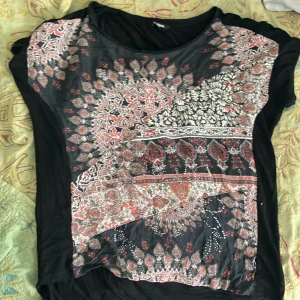 Svart topp med orientaliskt mönster - Svart topp från Desigual med ett detaljerat orientaliskt mönster i rött, vitt och beige och små stenar på. Tunt mjukt material. Passar S-M skulle jag säga, men står XL på.