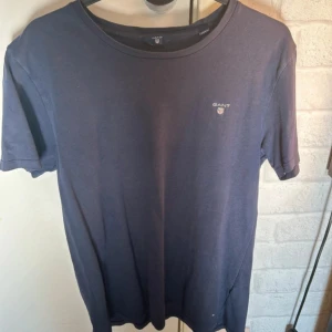 Mörkblå t-shirt från GANT - Snygg mörkblå t-shirt från GANT med klassisk rund hals och diskret broderad logga på bröstet. T-shirten är i mjuk bomull och har en normal passform med korta ärmar. Perfekt för dig som gillar stilrena och enkla plagg med en touch av premiumkänsla.