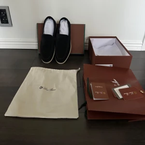 Svarta loafers från Loro Piana - Stilrena svarta loafers från Loro Piana i mjuk mocka med vit platt sula. Klassisk slip-on design med rund tå och diskret logga bak på höger häl. levereras med originalkartong, dustbag och allt annat som kommer med org🙌 Skambud besvaras ej👍pris kanske kan diskuteras vid smidig affär. Kan bytas mot maison margiela gats i storlek 44.
