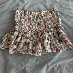 Blommig volangkjol i ljusa toner - Supersöt kort kjol med blommigt mönster i lila, rosa och svart på ljus botten. Kjolen har smock upptill och flera lager volanger som ger en härlig och lekfull look. Perfekt för dig som gillar romantisk stil och vill sticka ut lite extra.