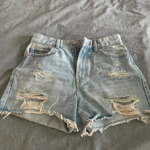 Ljusblå slitna jeansshorts - Säljer ett par ljusblå jeansshorts med hög midja och råa, fransiga kanter. Shortsen har flera slitningar och hål för en riktigt avslappnad och trendig look. Perfekta för sommardagar och festivaler.