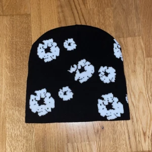 Svart mössa med vita blommor - Cool svart mössa med vita blommor i pixelmönster. Perfekt accessoar för att sticka ut i mängden och hålla värmen. Mjuk och stretchig, passar de flesta huvuden. Snygg detalj till din streetwear eller casual look.