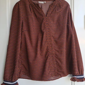 Mönstrad blus från Fransa i S - Säljer en snygg blus från Fransa i storlek S. Blusen har ett grafiskt mönster i mörkblått och orange, långa ärmar med volangdetalj och smal rand vid ärmslutet. Luftig passform och v-ringad hals. 