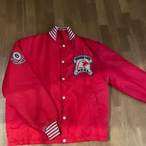 Röd bomberjacka Warren Park - Röd bomberjacka med vita ränder på krage och muddar. Jackan har broderade patches på bröstet och ena ärmen, samt knappar framtill. Perfekt för dig som vill sticka ut med en sportig och retro vibe.
