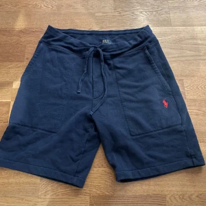 Mörkblå shorts från Polo Ralph Lauren - Mörkblå shorts från Polo Ralph Lauren med snörning i midjan och två stora fickor framtill. Klassisk röd logga broderad på ena benet. Sköna och avslappnade, perfekta för sommaren. Tillverkade i mjuk bomull för maximal komfort.