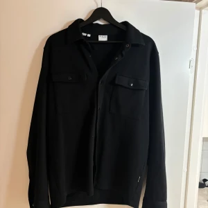 Svart overshirt från Selected Homme - Stilren svart overshirt från Selected Homme med klassisk krage, två bröstfickor med lock och tryckknappar. Långärmad modell med knappar vid ärmslut. Perfekt att bära öppen över en t-shirt för en avslappnad look. Ena knappen är borta vid vänster ärmslut så går ej att stäng igen vänster ärmslut, men funkar ändå att ha på sig 