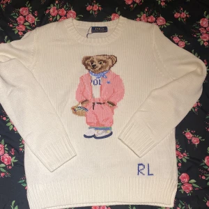 Vit stickad tröja Polo Bear - Vit stickad tröja från Polo Ralph Lauren med klassisk Polo Bear-motiv framtill. Björnen har rosa outfit och blå scarf, och RL-broderi vid nederkanten. Tröjan har rund hals och långa ärmar med ribbade muddar. Perfekt för dig som gillar preppy stil och ikoniska prints.