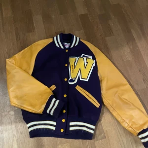 Gul och lila bomberjacka från DeLONG - Retro bomberjacka från DeLONG i lila med gula skinnärmar och stora W-patch på bröstet. Jackan har ribbade muddar, knappar framtill och två fickor. Perfekt för dig som vill sticka ut med en klassisk amerikansk college-look.