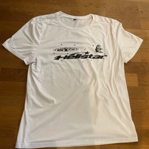 Vit Hellstar t-shirt med tryck - Snygg vit t-shirt från Hellstar i storlek M med svart grafiskt tryck på både fram- och baksidan. Framsidan har Hellstar-logga och flames, baksidan har ett stort motiv med profil och eldflames. T-shirten har rund hals och korta ärmar, perfekt för dig som gillar streetwear.