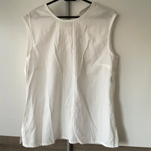 Vit ärmlös blus med broderi - Klassisk vit blus i bomull med ärmlös design och diskret broderad detalj framtill. 