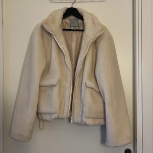 Vit/Beige teddyjacka från Urbancode - Supermjuk och fluffig teddyjacka från Urbancode London i vit/beige. Jackan har dragkedja framtill, två stora fickor och hög krage. Perfekt för kalla dagar när du vill vara både varm och snygg. Dragsko nertill för att justera passformen.