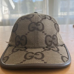 Gucci keps med GG-mönster beige - Snygg keps från Gucci i beige med det klassiska GG-mönstret i mörkbrunt. Kepsen har böjd skärm, justerbar rem bak i brunt skinn och är tillverkad i ett tåligt bomullsmaterial. Perfekt för dig som vill sticka ut med en lyxig accessoar.