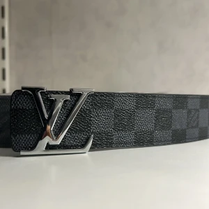 Louis Vuitton rutigt svart bälte - Snyggt svart bälte från Louis Vuitton med klassiskt rutigt mönster och en stor LV-logga i silver som spänne. Bältet är gjort i exklusivt skinn och har en modern, stilren look som sticker ut.