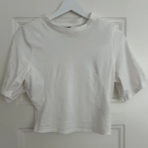 Vit croppad topp XS - Enkel och stilren vit croppad topp från Divided i storlek XS. Toppen har rund halsringning och korta ärmar som slutar strax ovanför armbågen. Tillverkad i mjuk bomull, perfekt till jeans eller kjol för en clean look.