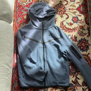 Blå Nike hoodiejacka XL med huva - Nike hoodiejacka i blått med hel dragkedja, huva och diskret logga på bröstet. Jackan har en praktisk ficka med dragkedja på ärmen och är tillverkad i mjukt material. Perfekt för chill dagar eller när du är på språng.